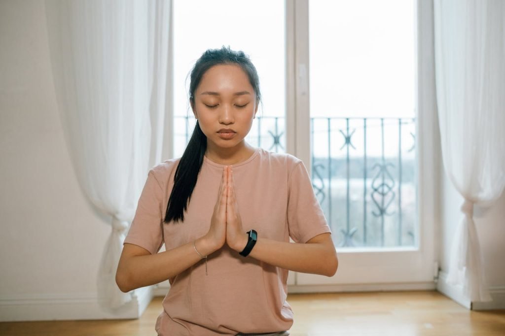 Asian woman practicing meditation indoors, hands together, promoting peace and healthy lifestyle.